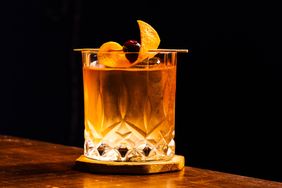 A cocktail in a crystal glass garnished with an orange peel and cherry placed on a wooden coaster