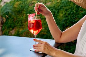 A person stirring a red beverage in a glass with a straw seated outdoors at a table
