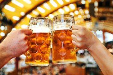 Two hands holding large beer steins in a celebratory gesture likely at an Oktoberfest event with a festive background