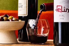 A glass being filled with red wine with bottles and a bowl of olives on a table
