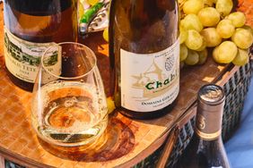 A picnic table scene with wine bottles a glass of white wine and grapes