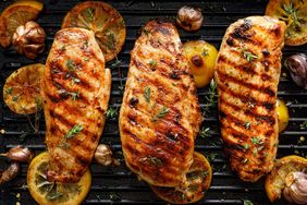 Three grilled chicken breasts on a grill with citrus slices and seasoning