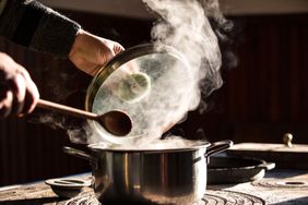 A persons hands lifting a pot lid while steam rises from the pot on a stove holding a wooden spoon