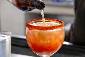 A beverage being prepared with a bottle pouring liquid into a rimmed glass filled with ice and garnished with reddish seasoning