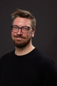 Headshot of Dylan Garret, smiling and facing forward, wearing black sweater and glasses on solid dark gray background