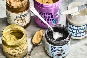 An assortment of open jars of spreadable foods including carrot cake pistachio and sesame butter placed on a countertop with utensils