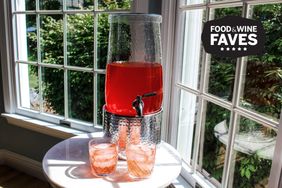 A drink dispenser with red liquid on a stand two glasses filled with the same drink placed on a table in front of windows