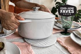 Hands placing a white ceramic pot with lid on a decorative trivet on a set dining table