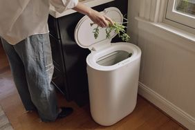 Person disposing vegetable scraps into a white compost bin with an open lid