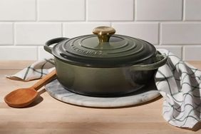 A round cast iron cooking pot with a lid on a circular marble surface next to a wooden spoon and a folded tea towel