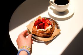 A hand reaching for a pastry topped with cream and strawberries on a plate beside a cup of coffee on a white table