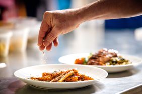 A persons hand sprinkling seasoning over cooked carrots on a plate another plate with food is in the background
