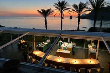 A restaurant with glass walls people inside preparing food & 168极速赛车官方开奖记录查询与实时直播overlooking a scenic coastline at sunset with palm trees and the sea