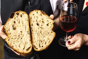 Two hands holding a cut loaf of bread and a glass of red wine respectively