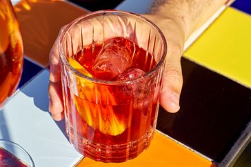A hand holding a glass of iced drink with orange slice on a colorful tiled surface