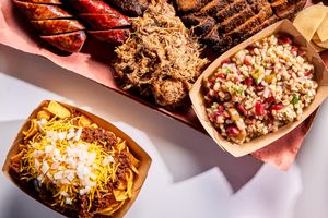 A platter of barbecue meats and side dishes including sausages brisket pulled pork a serving of corn salad and nachos with cheese and onions