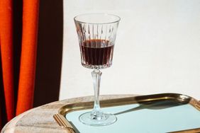 A cocktail glass with dark liquid on a table near a tray and a curtain