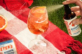 A glass with a beverage and a hand spraying Skeeters brand product nearby arranged on a red and white checkered cloth outdoors