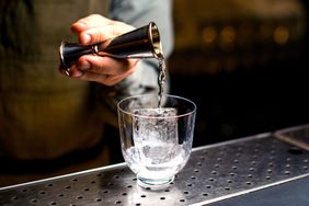 Pouring liquid from a jigger into a glass with ice cubes on a bar counter