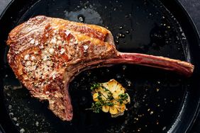A cooked tomahawk steak on a pan with roasted garlic and herbs