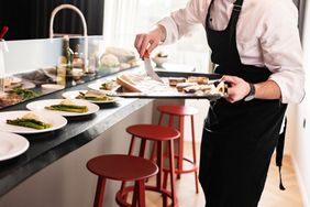 A chef preparing food in a kitchen plating dishes next to a counter with stools