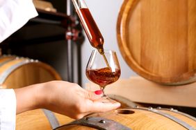 A persons hand holding a glass being filled with whiskey from a pipette against a background of wooden barrels