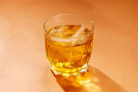 A rusty nail cocktail with a large block of ice on a peach colored surface. 