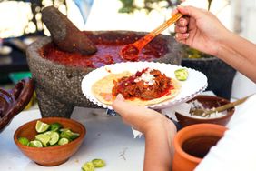 Person preparing a taco with spicy salsa from a traditional mortar surrounded by limes and bowls of condiments