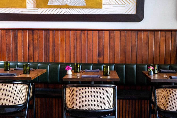 Interior of a restaurant with neatly set tables against a wooden panel wall featuring chairs with woven backs and small floral centerpieces
