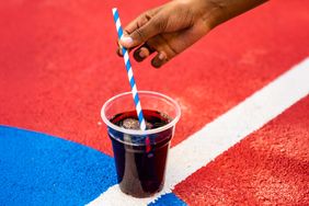 A hand holding a striped straw placed in a plastic cup with a dark beverage on a colorful surface with red blue and white stripes