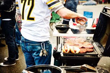 A person grilling hot dogs and patties on a portable grill during a tailgate gathering wearing a sports team jersey