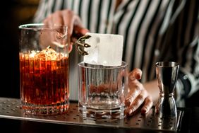 A bartender using tongs to place a large ice cube into a cocktail glass next to a mixing glass and a jigger