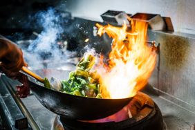 Chef stir-frying vegetables in a wok with a visible flame on the stovetop