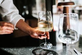 A hand touching a glass of white wine on a table with another glass and cutlery nearby indoor setting visible in the background