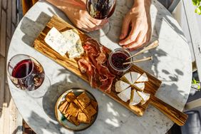 A gourmet cheese and charcuterie platter with wine on a table hands visible reaching for food