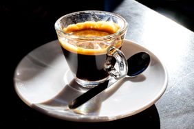A glass cup of espresso on a white saucer next to a spoon sitting on a table under bright lighting