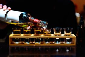 A person pouring liquor into a row of shot glasses on a wooden tray